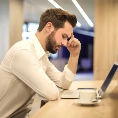 hombre trabajando en mesa con portatil y cafe con la mano sobre la frente