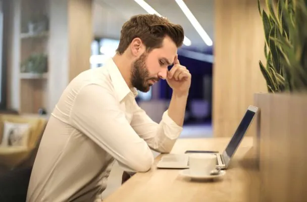hombre trabajando en mesa con portatil y cafe con la mano sobre la frente